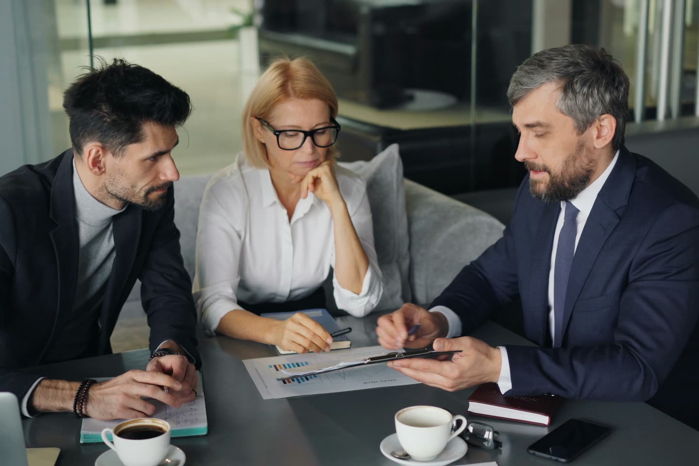 Três profissionais sentados à mesa analisando um relatório com gráficos, com café e anotações