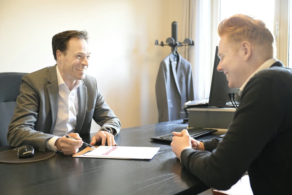 Profissional sorrindo enquanto preenche um documento com um cliente à mesa